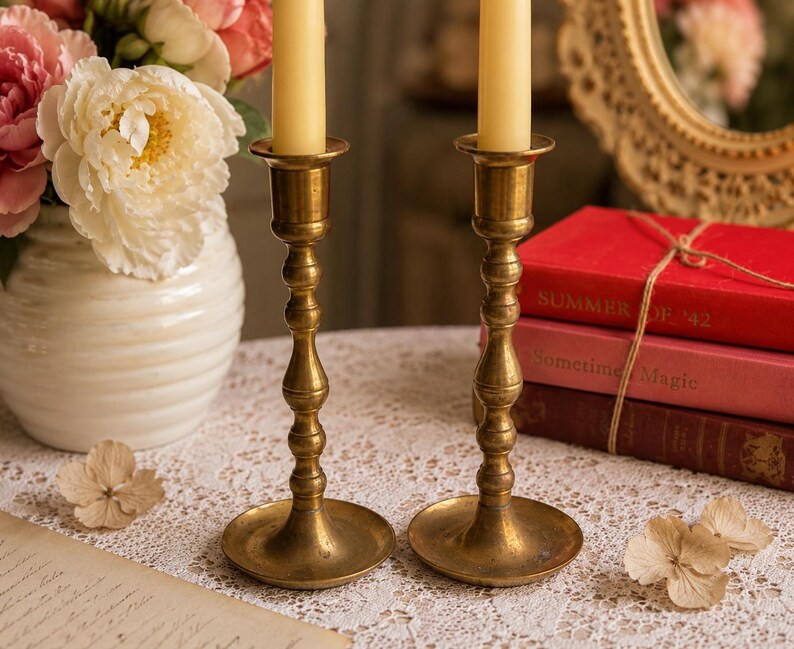 May include: Two matching brass candlesticks with a polished finish and a tiered, slender stem design. Each candlestick holds a single cream-colored taper candle. The brass metal has a warm, golden tone with a smooth, reflective surface texture.