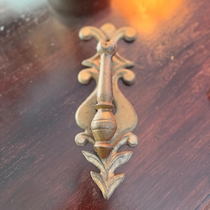 May include: A vintage brass drawer pull with an ornate design. The pull features a central handle with a bulbous midsection, flanked by decorative flourishes and a stylized arrow-shaped base. The brass has a warm, aged patina.