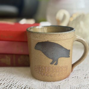 May include: A tan ceramic mug with a dark gray bird silhouette and a textured pattern at the base. The mug has a handle and a slightly speckled finish, suggesting a handmade quality. The mug is on a white lace tablecloth.