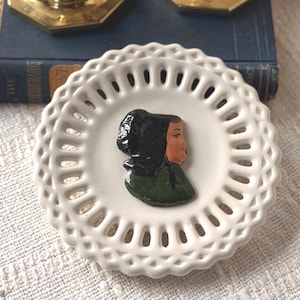 May include: A white, scalloped-edge ceramic plate with a decorative relief of a woman's profile. The woman's head is covered with a black headscarf, and she wears a green garment. The plate has a series of small, evenly spaced holes around the edge.