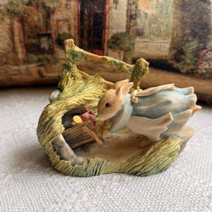 May include: A ceramic figurine of a brown rabbit wearing a light blue jacket, standing next to a small wooden wheelbarrow filled with vegetables. The rabbit is positioned near a textured, grassy mound and a small wooden fence structure.