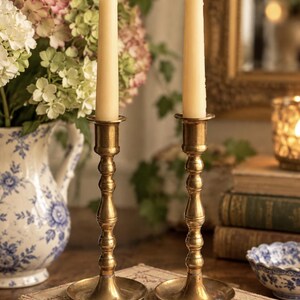 May include: Two matching brass candlesticks with a slender, tiered stem design and a wide circular base. Each candlestick holds a single, tall, smooth, cream-colored taper candle. The metal has a polished, warm, metallic gold-toned finish.