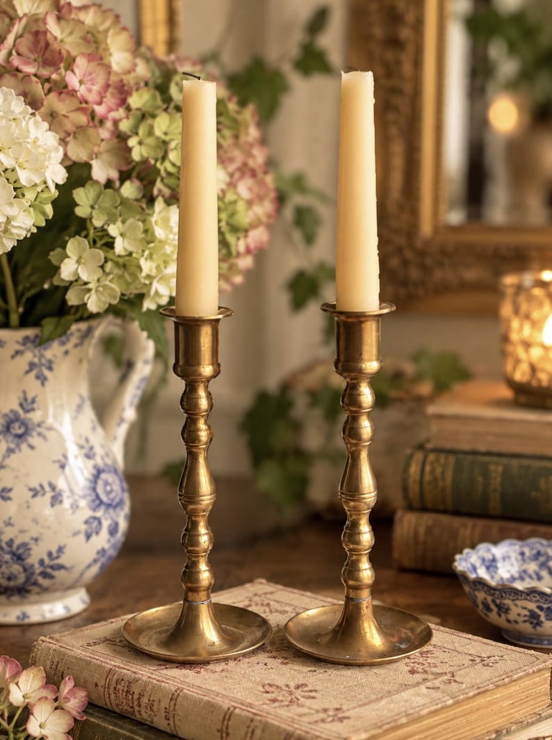 May include: Two matching brass candlesticks with a slender, tiered stem design and a wide circular base. Each candlestick holds a single, tall, smooth, cream-colored taper candle. The metal has a polished, warm, metallic gold-toned finish.