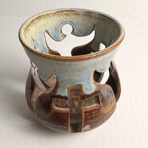 May include: A ceramic candle holder with a light blue and brown glaze. The holder features cut-out designs of figures and a cross. The top rim is light blue, and the base is brown. The candle holder is a decorative item.