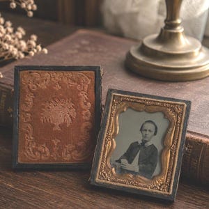 May include: Two rectangular antique daguerreotype cases. One case is closed, featuring a brown velvet cover with an embossed floral urn design. The other case is open, revealing a gold-toned metal frame containing a black and white portrait photograph.
