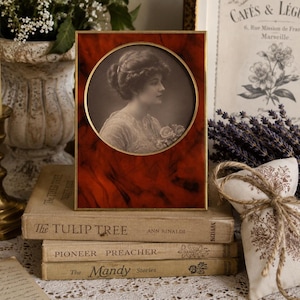 May include: A rectangular picture frame with a deep red, marbled finish and a thin gold-toned metal border. The frame features a circular central opening containing a black and white portrait of a person with an updo hairstyle holding flowers.