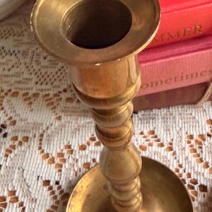 May include: A polished brass candlestick holder featuring a wide circular base, a slender stem with multiple decorative rounded nodes, and a flared top opening designed to securely hold a single taper candle in an upright position.