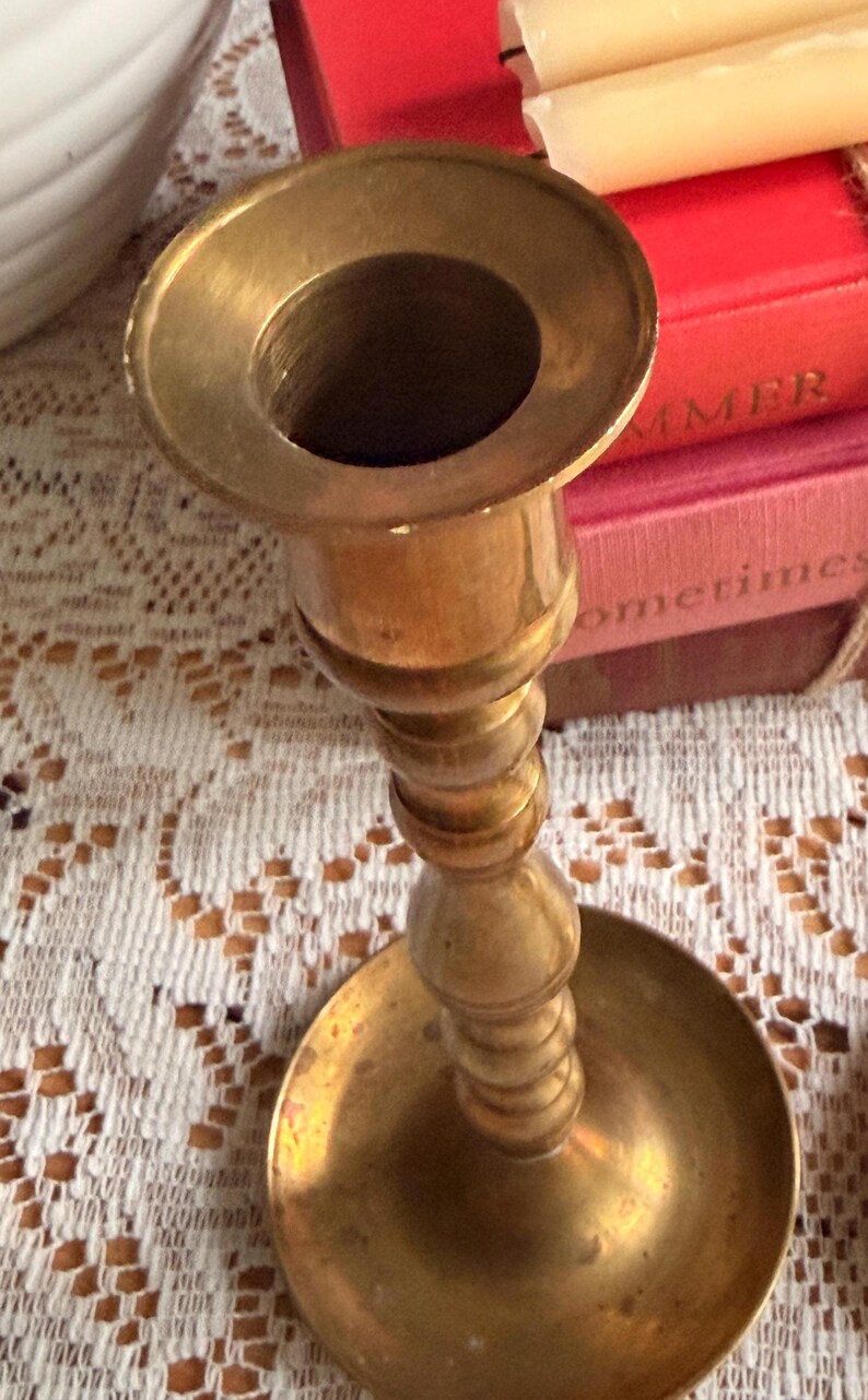 May include: A polished brass candlestick holder featuring a wide circular base, a slender stem with multiple decorative rounded nodes, and a flared top opening designed to securely hold a single taper candle in an upright position.