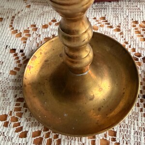 May include: A brass candlestick holder featuring a wide, circular base and a central stem with rounded, tiered sections. The metal surface displays a warm, golden-yellow tone with subtle variations in color and texture across the circular tray.