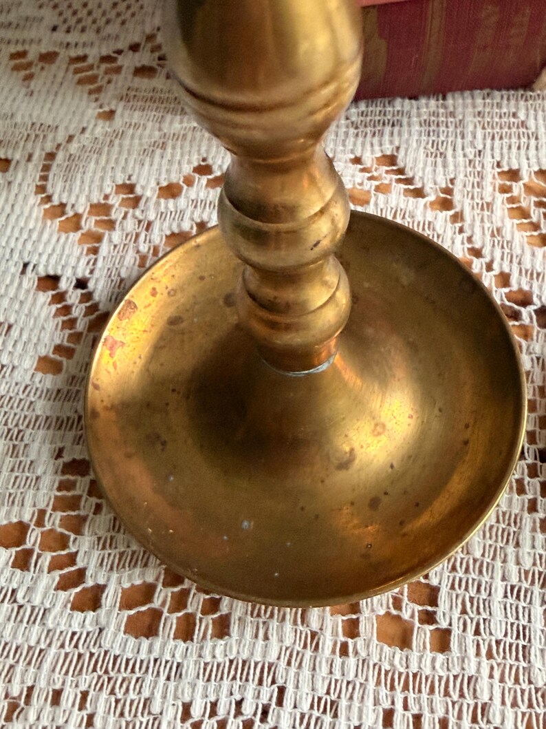May include: A brass candlestick holder featuring a wide, circular base and a central stem with rounded, tiered sections. The metal surface displays a warm, golden-yellow tone with subtle variations in color and texture across the circular tray.