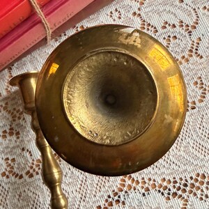 May include: A top-down view of a polished brass candlestick holder with a circular base and a slender, multi-tiered stem. The metal surface shows a warm, reflective golden tone with subtle signs of wear and natural patina on the base.