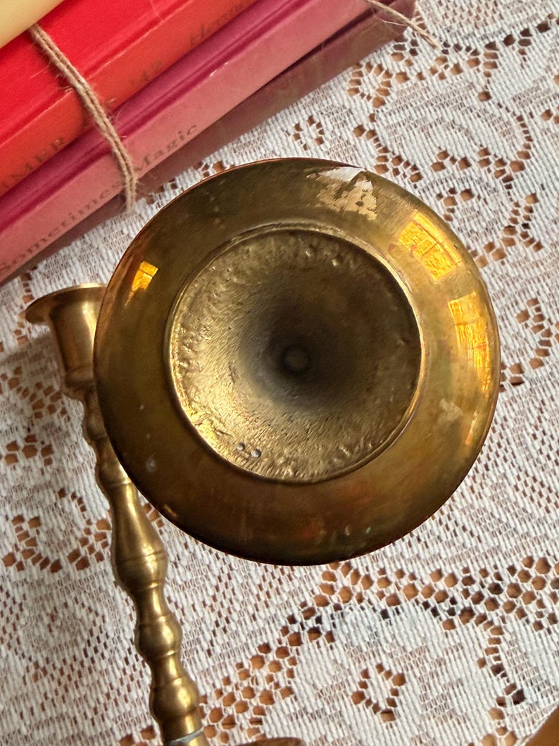 May include: A top-down view of a polished brass candlestick holder with a circular base and a slender, multi-tiered stem. The metal surface shows a warm, reflective golden tone with subtle signs of wear and natural patina on the base.