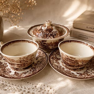 May include: A vintage tea set with a brown and white landscape pattern. The set includes two teacups with saucers and a covered serving dish. The teacups and saucers are placed on a white surface with a floral design.