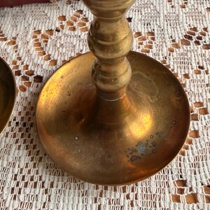 May include: A brass candlestick holder featuring a wide, circular base with a polished metallic finish and visible signs of natural oxidation. The central stem displays a series of rounded, stacked decorative rings leading upward from the base.
