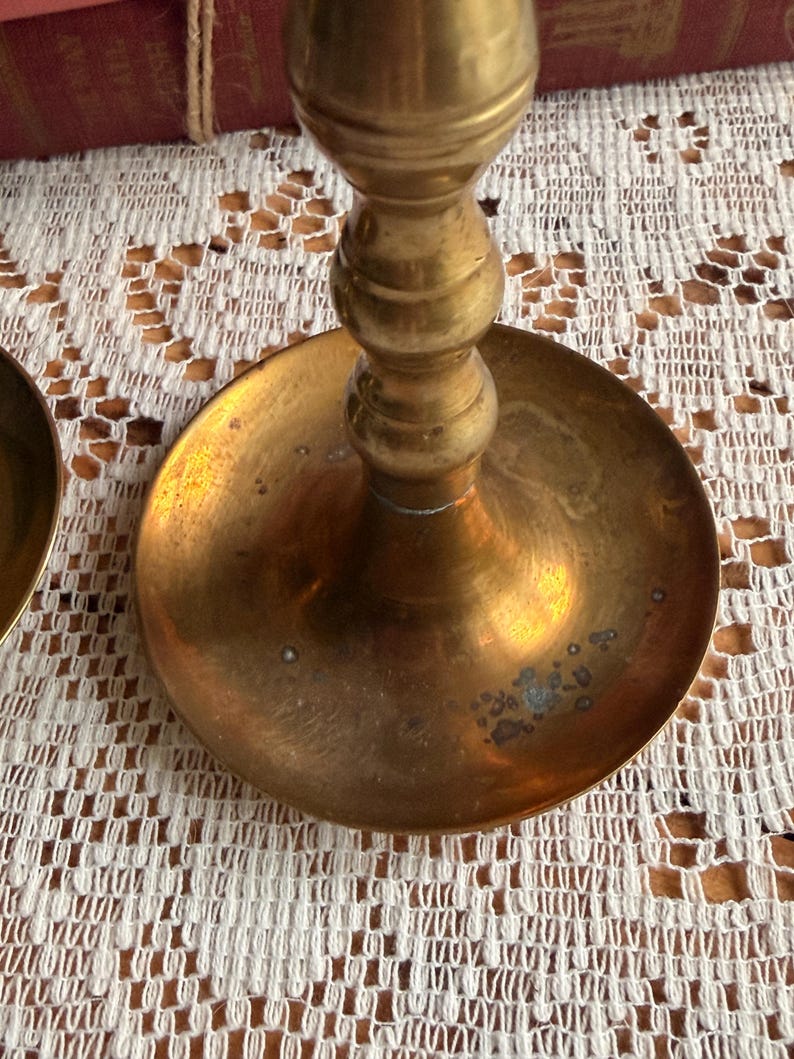 May include: A brass candlestick holder featuring a wide, circular base with a polished metallic finish and visible signs of natural oxidation. The central stem displays a series of rounded, stacked decorative rings leading upward from the base.