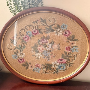 May include: An oval wall hanging featuring a needlepoint floral design with a central bouquet and a surrounding wreath of pink, blue, and white flowers on a tan background, encased in a dark brown wooden frame with a thin gold inner trim.