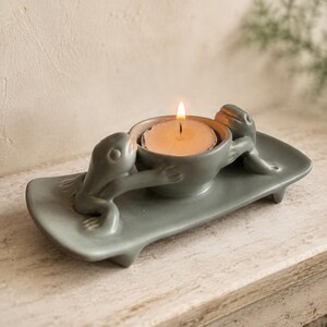 May include: A matte sage green ceramic candle holder featuring two sculpted frogs positioned on opposite sides of a central circular bowl. The bowl contains a single lit white tea light candle, casting a warm glow over the smooth, curved surface.