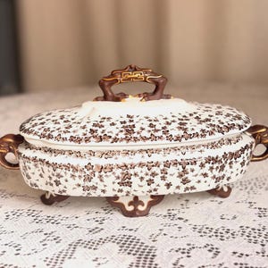 May include: Oval, lidded serving dish with a white background and a brown floral pattern. The dish has brown handles and feet. The lid has a decorative brown handle. The dish is on a white lace tablecloth.