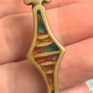 May include: A vintage gold-toned pendant with a colorful enamel inlay. The pendant has a unique shape with a diamond-like center filled with orange, green, and yellow enamel. A small green gem is set at the bottom. The pendant is held by a hand.