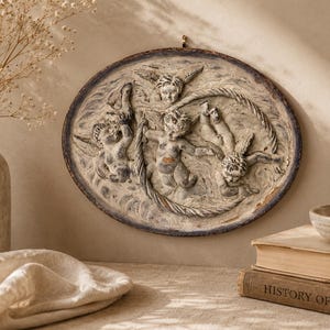 May include: A circular, antique-style relief sculpture featuring cherubic figures. The artwork is in shades of gray and beige, with a weathered appearance. The cherubs are arranged in a circular pattern, with a textured, aged finish.