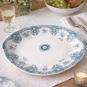 May include: An oval, white ceramic platter with a blue floral design around the rim and center. The platter is on a white tablecloth, with a glass of wine, flowers, and food in the background. The plate is approximately 14 inches long.