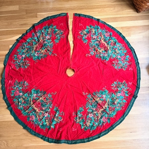 May include: A circular red Christmas tree skirt featuring a dark green border and a repeating pattern of brown deer, green holly leaves, red berries, and pinecones arranged in a decorative wreath-like design around the center opening.