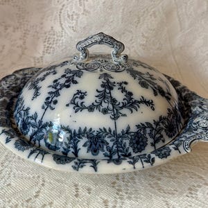 May include: A white and blue ceramic butter dish with a lid. The dish features a floral pattern in blue against a white background. The lid has a handle and the dish has a scalloped edge.
