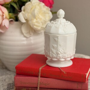 May include: A white, lidded, decorative jar with a detailed grape and leaf design, sitting atop a stack of three vintage books tied with twine. The books are various shades of red and pink. A white vase with pink and white flowers is in the background.