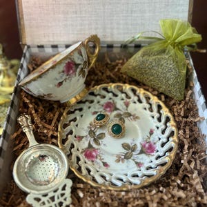 May include: A vintage-style gift set featuring a teacup, saucer, and tea infuser. The teacup and saucer have a floral design with gold trim. A small green bag of tea and a pair of green stone earrings are also included.