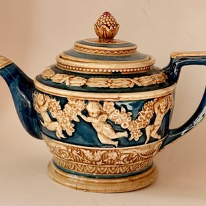 May include: A ceramic teapot with a blue and cream color scheme. The teapot features a detailed relief design with cherubs, floral elements, and a decorative lid with a finial. The handle and spout are blue, and the teapot has a classic, elegant shape.
