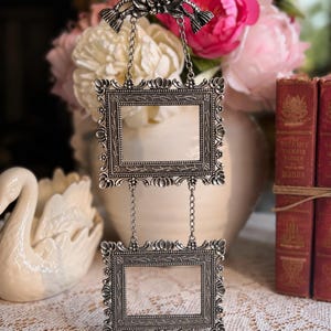 May include: A silver-toned metal hanging frame featuring two rectangular openings connected by delicate chains. The top piece has a decorative bow and floral motif, while both frames are detailed with intricate embossed patterns and scalloped edges.
