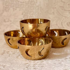 Pode incluir: Quatro pequenas tigelas redondas de latão com recortes de meia-lua. As tigelas são empilhadas e dispostas sobre uma superfície branca texturizada. As tigelas têm um acabamento dourado brilhante e estão vazias.