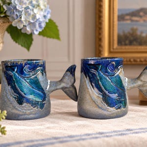 May include: Two ceramic mugs featuring a raised, sculpted whale design in shades of deep blue and teal. The mugs have a textured, speckled grey base and a handle shaped like a whale tail. The glaze is glossy on the whale and matte on the base.