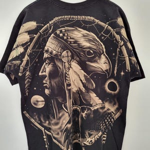May include: Black t-shirt featuring a Native American profile with an eagle head, feathers, and a dreamcatcher design. The artwork is in shades of brown and tan against a black background. The shirt is hanging on a wooden hanger.
