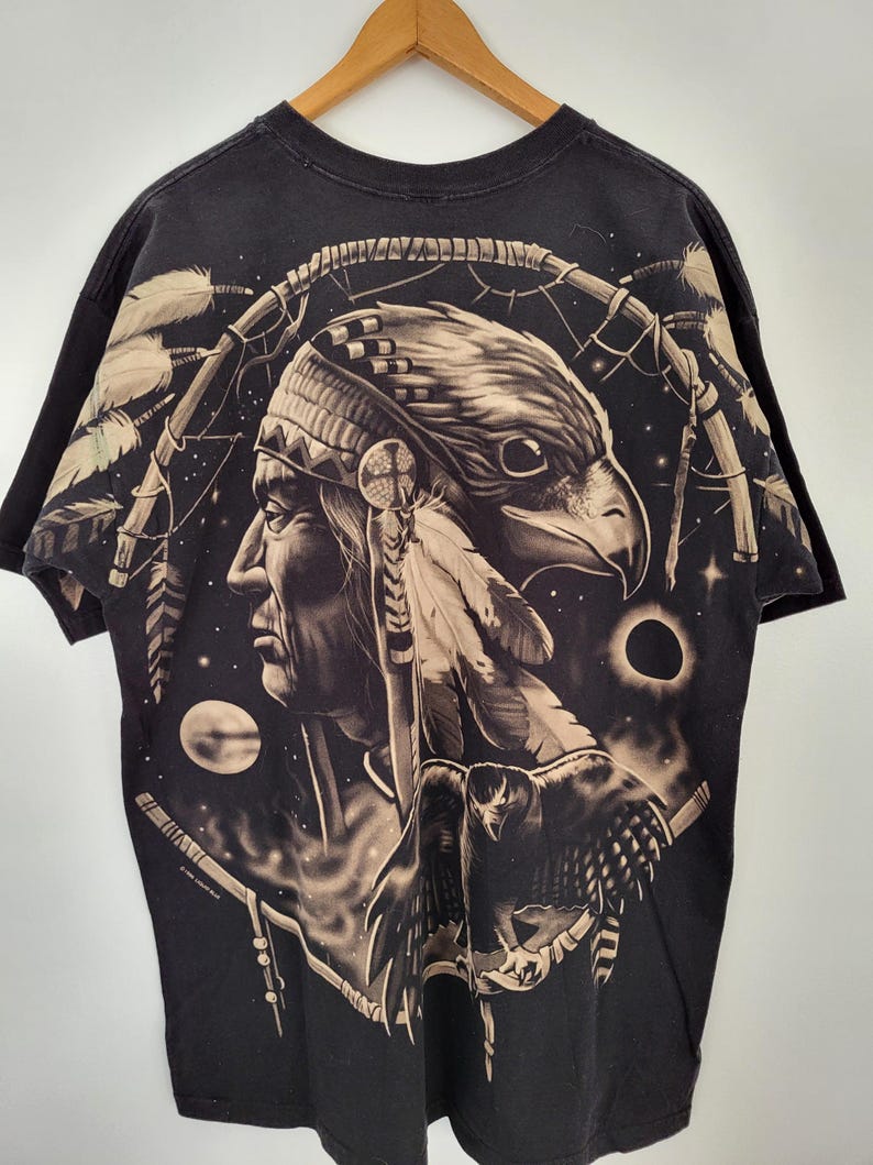 May include: Black t-shirt featuring a Native American profile with an eagle head, feathers, and a dreamcatcher design. The artwork is in shades of brown and tan against a black background. The shirt is hanging on a wooden hanger.