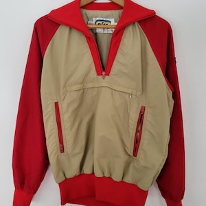 May include: A vintage anorak jacket in red and tan. The jacket features a red collar, sleeves, and cuffs, with a tan body and a half-zip closure. Two red zippered pockets are on the front. The jacket is hanging on a wooden hanger.