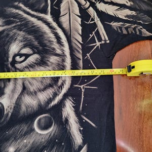 May include: Black t-shirt featuring a detailed wolf graphic with a dreamcatcher and feathers. The design is in shades of gray and white, with a celestial background. The tape measure shows the shirt is approximately 25 inches wide.