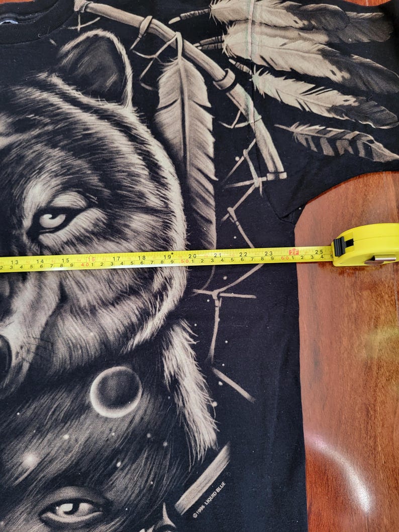 May include: Black t-shirt featuring a detailed wolf graphic with a dreamcatcher and feathers. The design is in shades of gray and white, with a celestial background. The tape measure shows the shirt is approximately 25 inches wide.