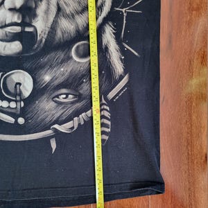 May include: Black t-shirt featuring a detailed graphic of a wolf's face and a human face, with a celestial design. The shirt is being measured with a yellow tape measure. The graphic is in shades of gray and white.