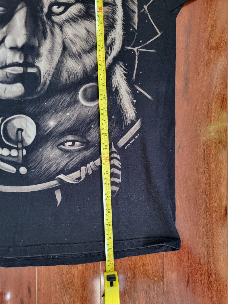 May include: Black t-shirt featuring a detailed graphic of a wolf's face and a human face, with a celestial design. The shirt is being measured with a yellow tape measure. The graphic is in shades of gray and white.