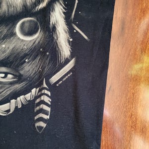 May include: Black t-shirt featuring a detailed illustration of a wolf's face with a moon and stars. The design is in shades of gray and white, with the text "&copy;1996 LIQUID BLUE" printed on the lower right side.
