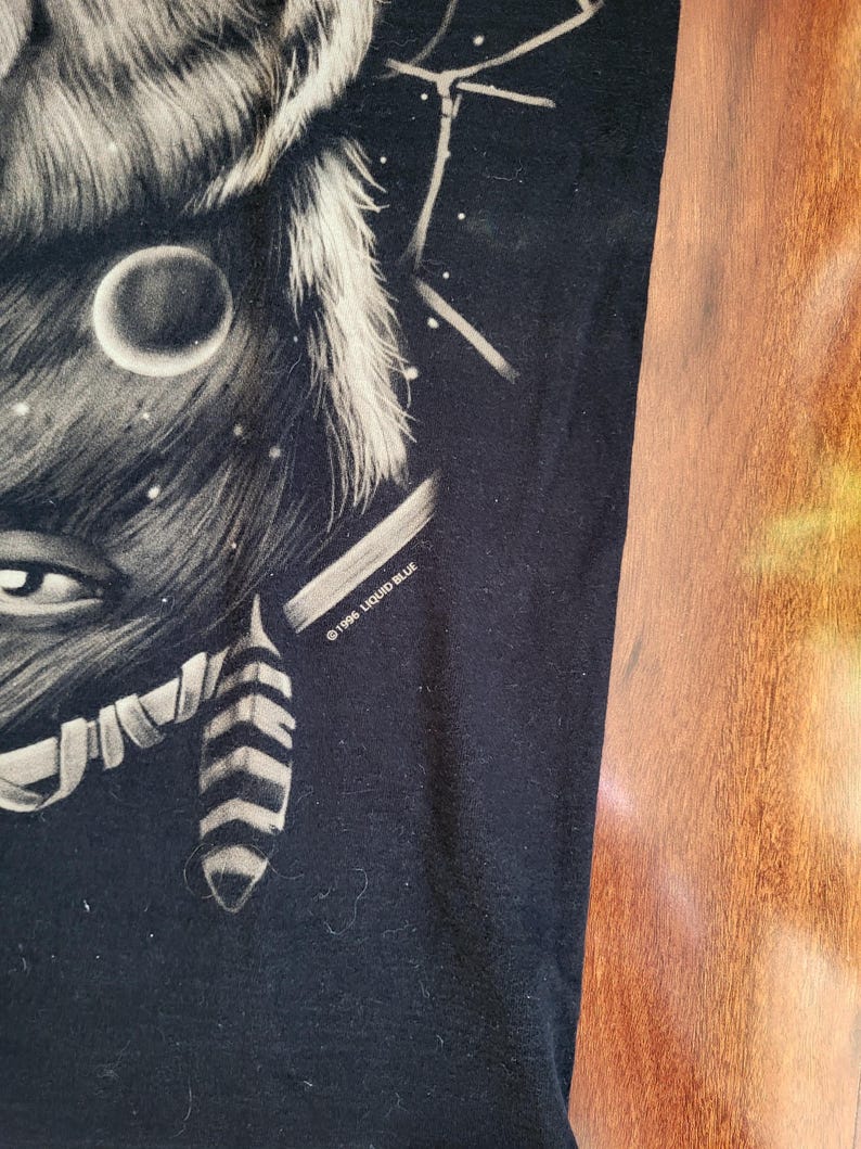 May include: Black t-shirt featuring a detailed illustration of a wolf's face with a moon and stars. The design is in shades of gray and white, with the text "&copy;1996 LIQUID BLUE" printed on the lower right side.