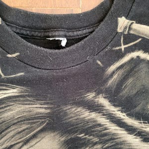 May include: Close-up of a black t-shirt featuring a detailed, faded graphic. The design appears to be a stylized image of a wolf or similar animal. The shirt's collar is visible, showing a ribbed texture.