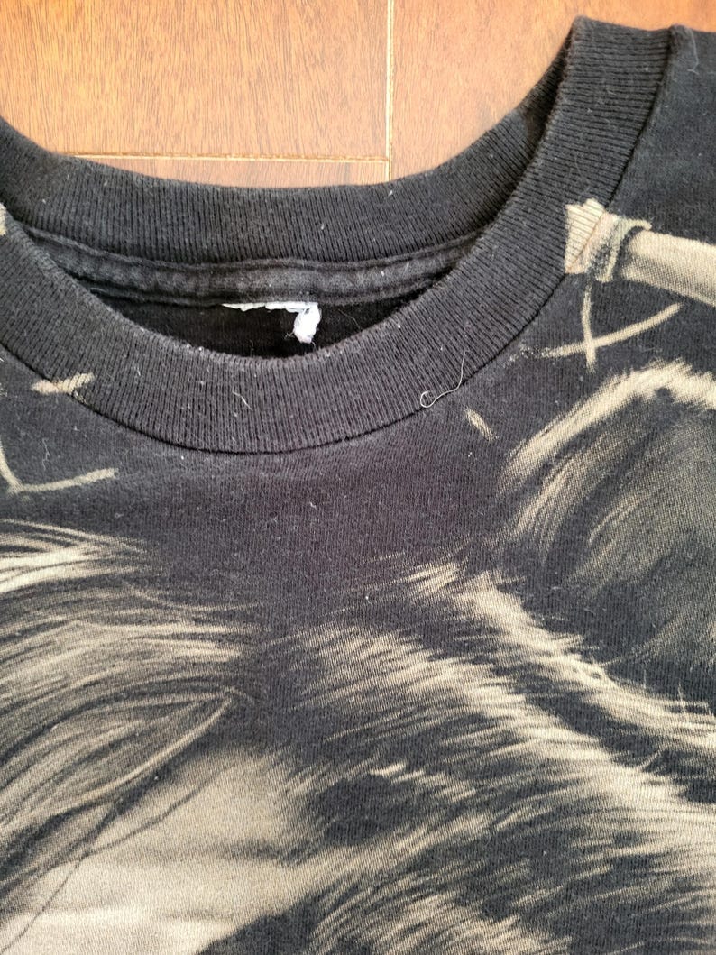 May include: Close-up of a black t-shirt featuring a detailed, faded graphic. The design appears to be a stylized image of a wolf or similar animal. The shirt's collar is visible, showing a ribbed texture.