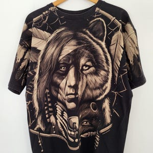 May include: Black t-shirt featuring a detailed graphic of a Native American face merged with a bear's head. The design incorporates feathers and tribal elements in shades of tan and brown. The shirt is hanging on a wooden hanger.