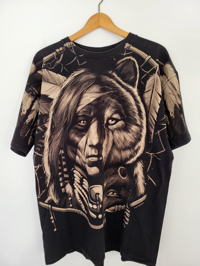 May include: Black t-shirt featuring a detailed graphic of a Native American face merged with a bear's head. The design incorporates feathers and tribal elements in shades of tan and brown. The shirt is hanging on a wooden hanger.