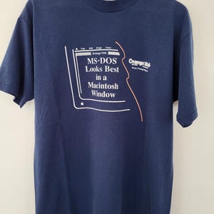 May include: Navy blue t-shirt with a white graphic of a Macintosh computer and the text "MS-DOS Looks Best in a Macintosh Window." The shirt also features the text "Orange 186."