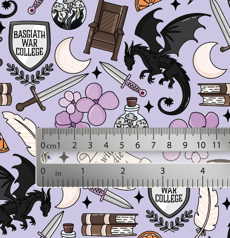 May include: A light purple fabric featuring a repeating pattern of black dragons, swords, shields labeled Basgiath War College, books, moons, and flowers. A metal ruler is placed across the center, showing measurements in inches and centimeters.