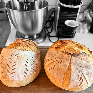 Könnte beinhalten: Zwei frisch gebackene Sauerteigbrote mit einem dekorativen Blattmuster auf der Oberseite. Das Brot liegt auf einem Holzbrett.