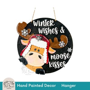 May include: A round, hand-painted wooden sign with a black background and the words "Winter wishes & moose kisses" in white. It features a cartoon moose wearing a red and white hat and snowflake accents. A decorative hanger.